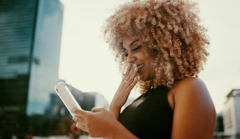 A woman in a black tank top smiles and covers her mouth as she looks down at her phone. This photo is being used in an article about dating profiles.