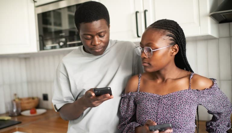 A young couple stands in the kitchen, leaning against the counter as they look down at their phones. The woman appears to be looking over at the man