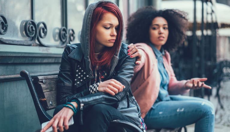 Two women sit outside on a metal bench. The woman in the foreground has her back turned to the woman beside her and appears to be avoiding her. The woman in the background has her hand on her friend