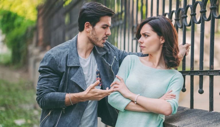 A woman leans against a fence, away from a man who has his arm behind her and is trying to talk to her. She looks visibly upset and has her arms crossed over her chest. This photo is being used in an article about bristle reactions in relationships.