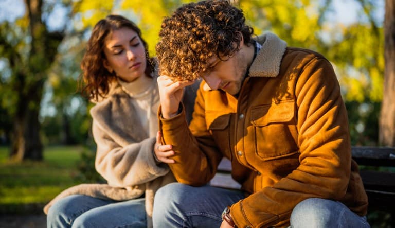 A young couple is sitting in the park on sunny day. The man is sad and the woman in consoling him. This photo is being used in an article about relationships fizzling out.