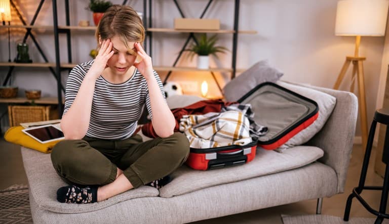 A woman sits cross-legged on a gray chaise next to an open suitcase. She is touching her temples with both hands and appears to be struggling with a headache. This photo is being used in an article about being tired after vacation.