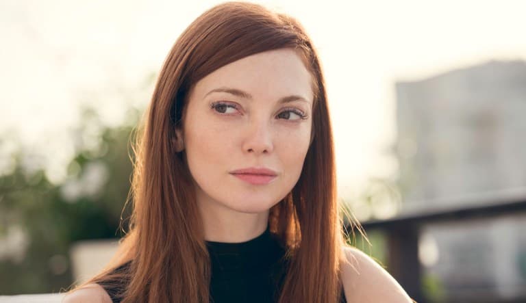 A woman with red hair stands outside looking off to the side. She is wearing a black, sleeveless shirt and looks contemplative. This photo is being used in an article about signs of an untrustworthy person.